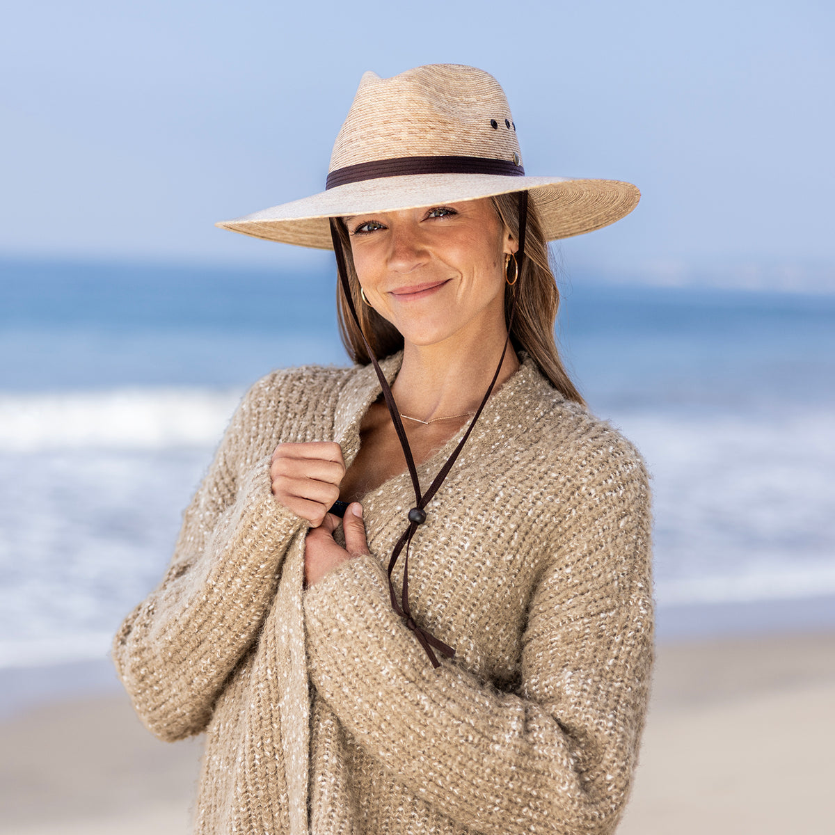 A woman standing on a beach wearing the Baja UPF 50+ palm fiber hat in camel with a chinstrap and a chunky knit beige cardigan.