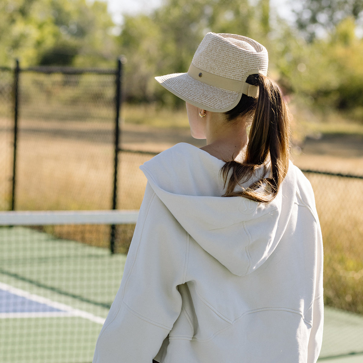 Woman playing tennis wearing the Wallaroo Women's Gabi Wide Brim Ponytail Fedora Sun Hat, featuring UPF 50+ protection, a ponytail opening, and a packable design for outdoor activities, Beige