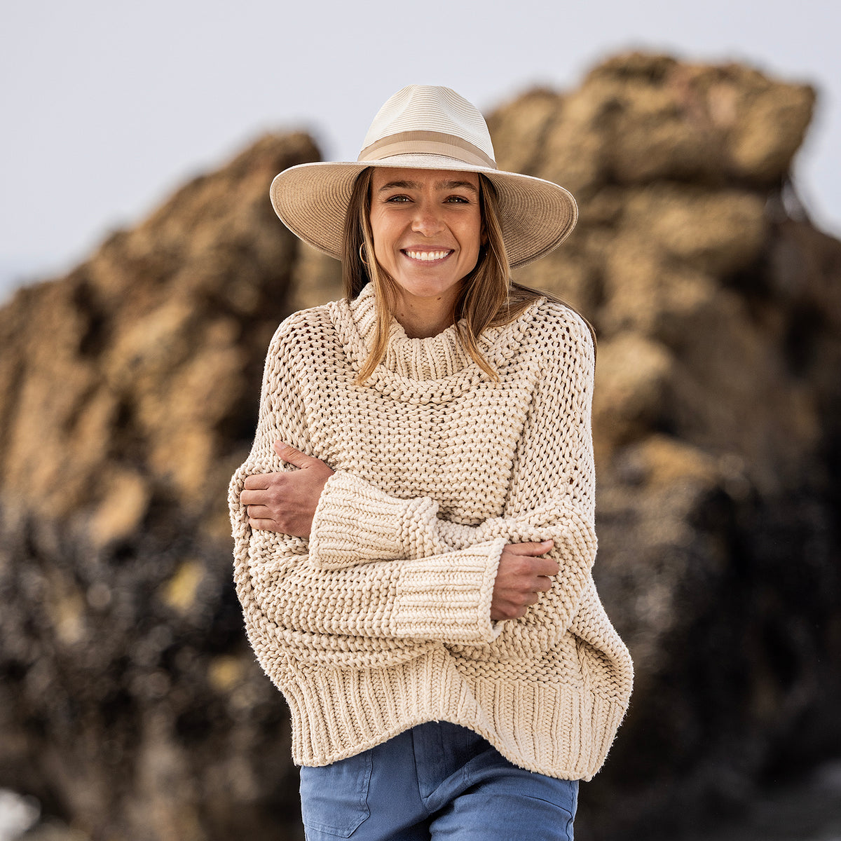 A smiling woman outdoors wearing the Wallaroo Solitaire hat by Jane Seymour in mixed camel, a beige knit sweater, and blue pants against a blurred natural rock background.