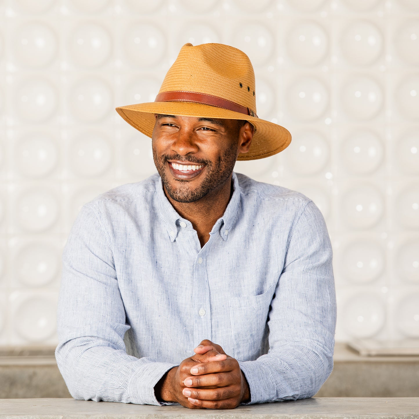 Man wearing the Avery Japanese paper glaze sun hat and light blue shirt against a textured wall, Camel