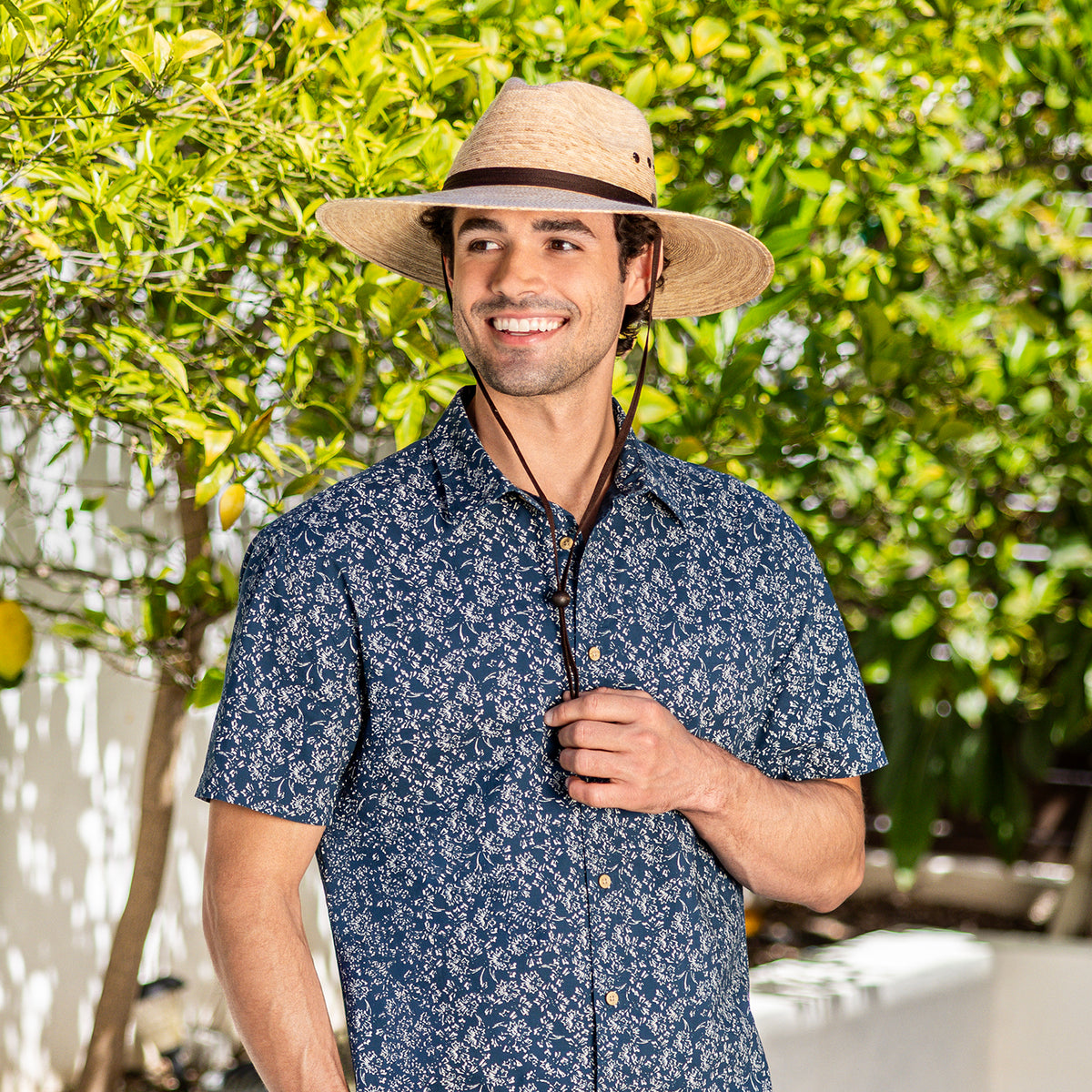 A man wearing a blue and white patterned short-sleeve button-up shirt and the Baja UPF 50+ palm fiber hat in camel with a chinstrap, standing outdoors in front of green foliage.