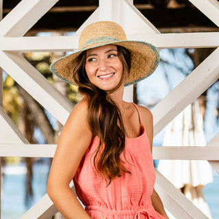 Woman wearing the Wallaroo Women's Turquoise Camille Raffia Sun Hat with a wide brim, providing packable, eco-friendly sun protection for travel, sightseeing, and summer outings