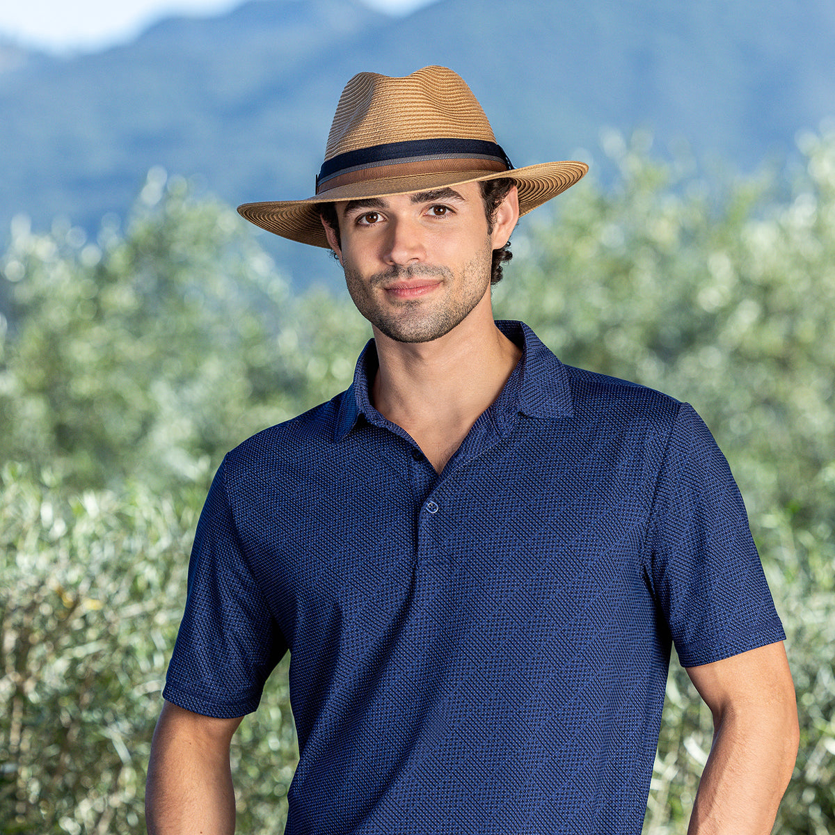 A man wearing the Carkella Colin fedora hat in camel and a textured navy blue short-sleeve polo shirt. He is standing outdoors against a blurred background of green vegetation and distant mountains under a clear sky