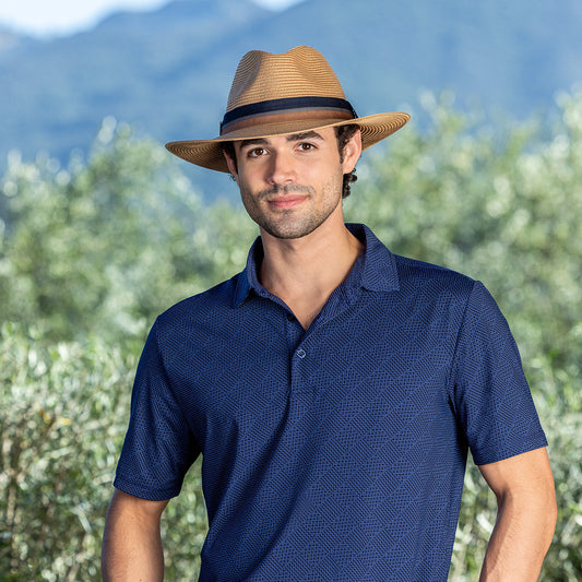 A man wearing the Carkella Colin fedora hat in camel and a textured navy blue short-sleeve polo shirt. He is standing outdoors against a blurred background of green vegetation and distant mountains under a clear sky