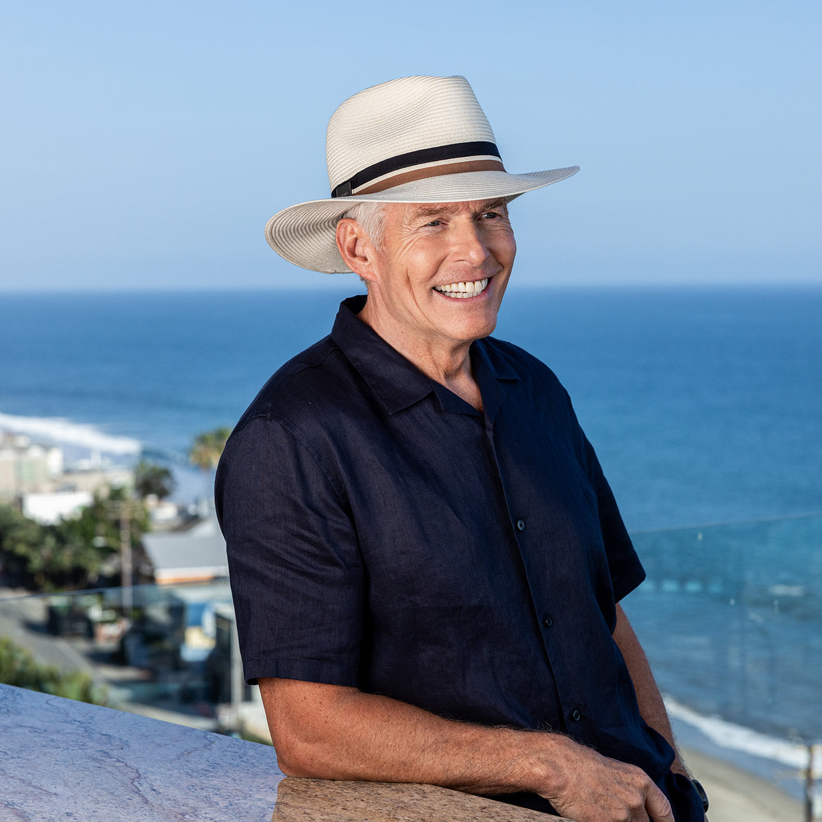 A smiling man wearing the unisex Carkella Colin fedora hat in ivory, paired with a short-sleeve dark blue button-down shirt, standing outdoors with an ocean view in the background.