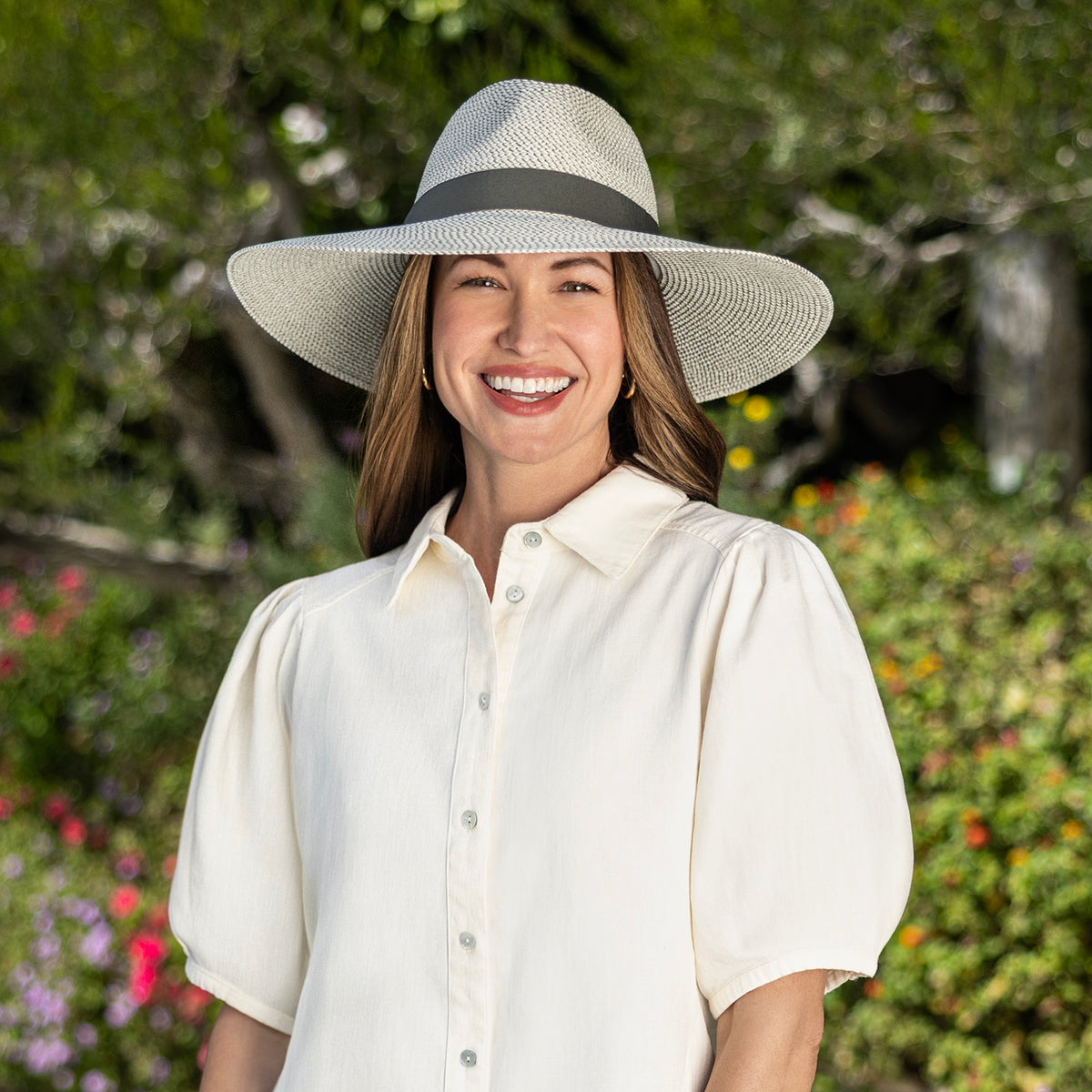 A smiling woman wearing the wide-brimmed Carkella Kerrigan UPF 50+ fedora hat in ivory olive with a dark green band and a cream puff-sleeve button-up shirt, photographed outdoors against a blurred garden background.