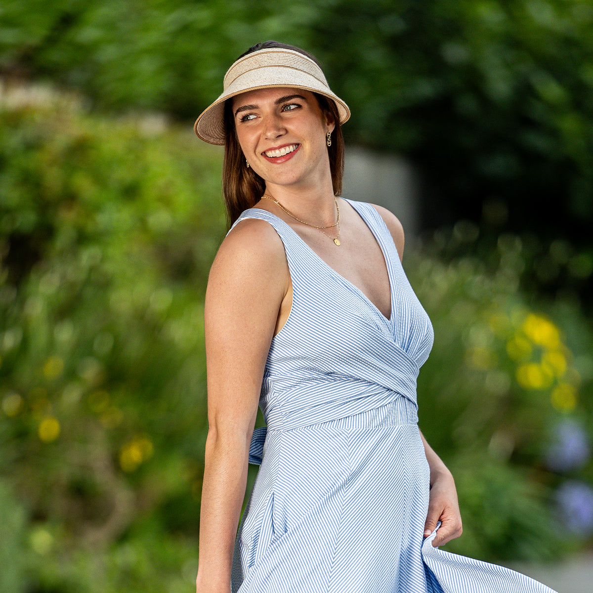 A woman wearing a light blue and white striped wrap-style midi dress and the packable Carkella Lily Visor in white/beige, smiling outdoors against a blurred green garden background.