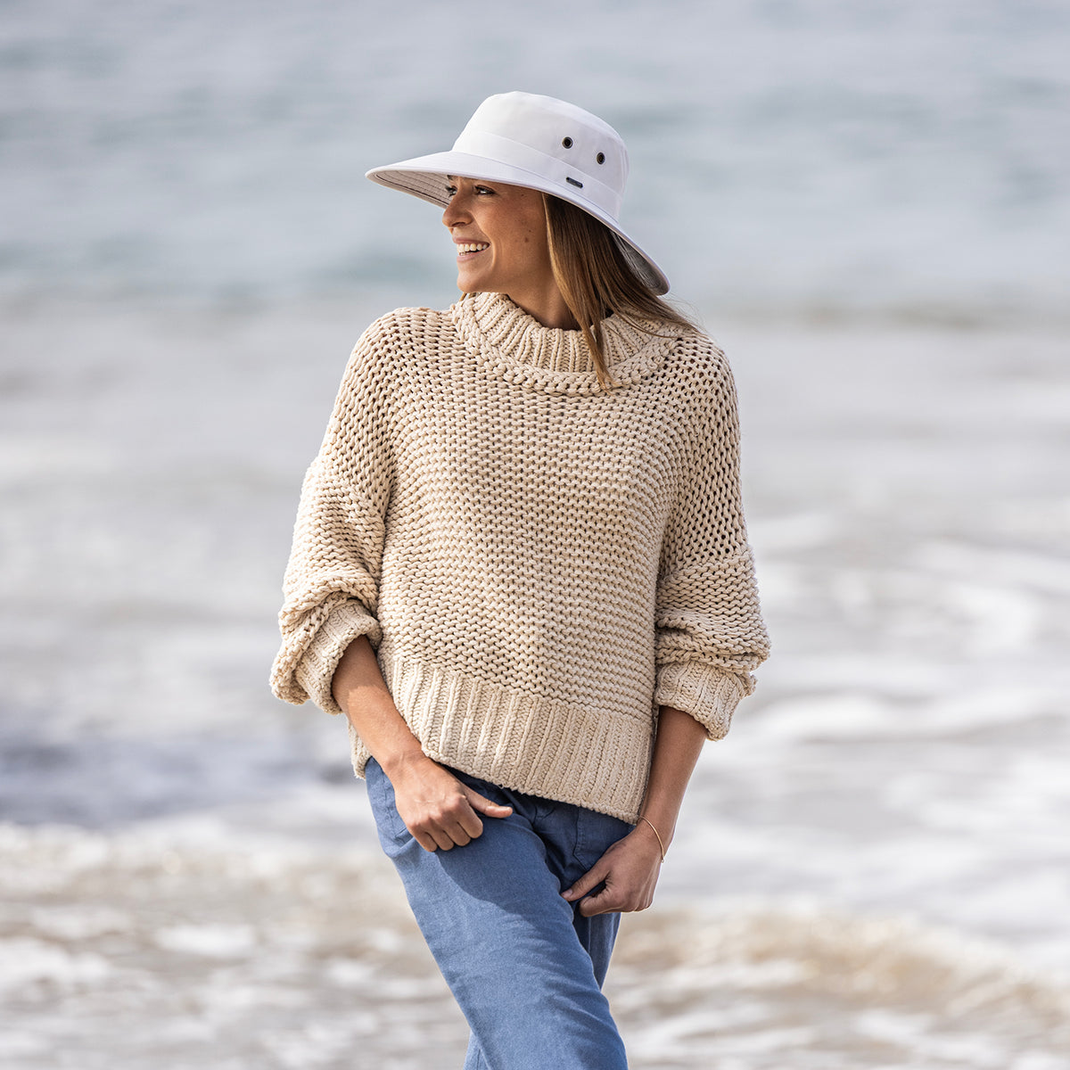 A woman wearing a chunky cream knit mock-neck sweater, blue jeans, and the Carkella Tahoe UPF 50+ packable hat in white standing on a beach with the ocean in the background.