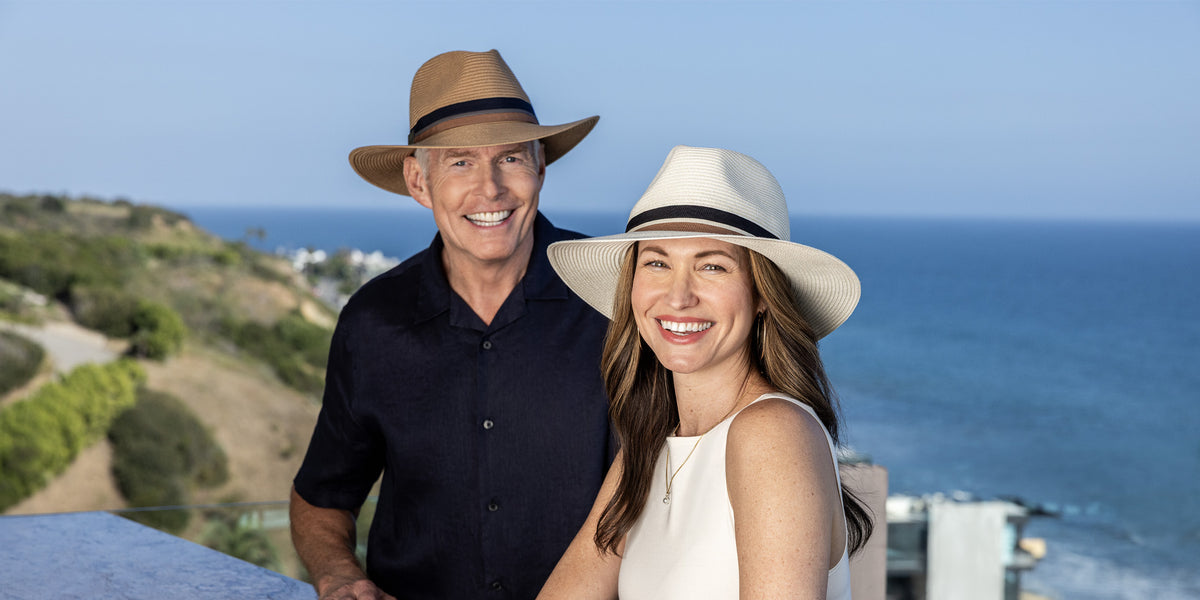 A smiling man and woman standing outdoors near the ocean, with the man wearing the unisex Carkella Colin fedora hat in camel and the woman wearing the same unisex style in ivory.