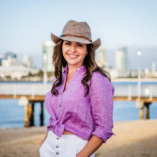 Woman wearing the Wallaroo Catalina Cowboy Crocheted Raffia Sun Hat while enjoying a sunny beach walk, offering stylish sun protection with a wide brim and perfect for women with larger head sizes, Mushroom