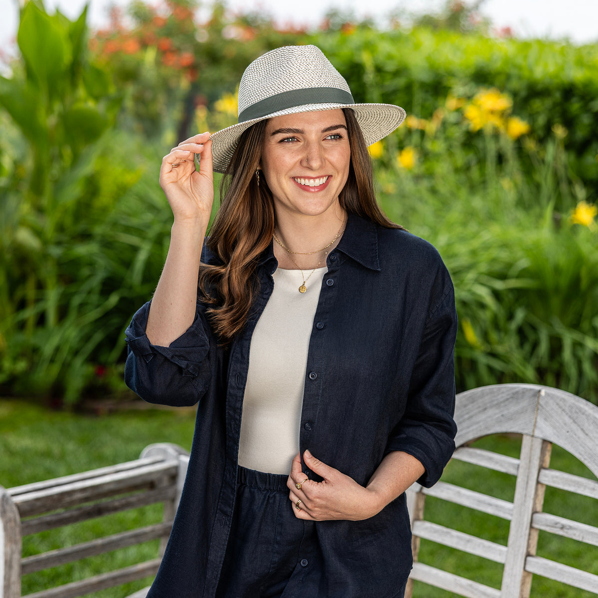 A woman wearing a dark navy button-up shirt, a cream top, and the Charlie UPF 50+ hat in olive, standing in a sunny garden next to a white wooden bench.