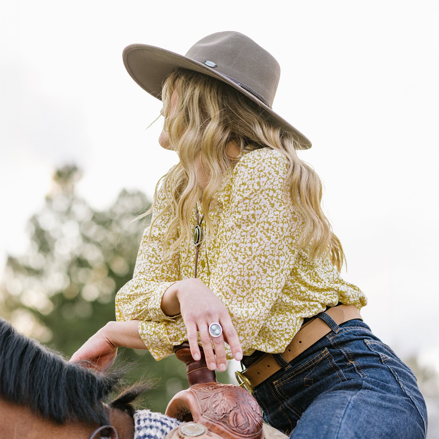 Woman in a yellow floral shirt and Colorado winter felt hat riding a horse, with a natural background.