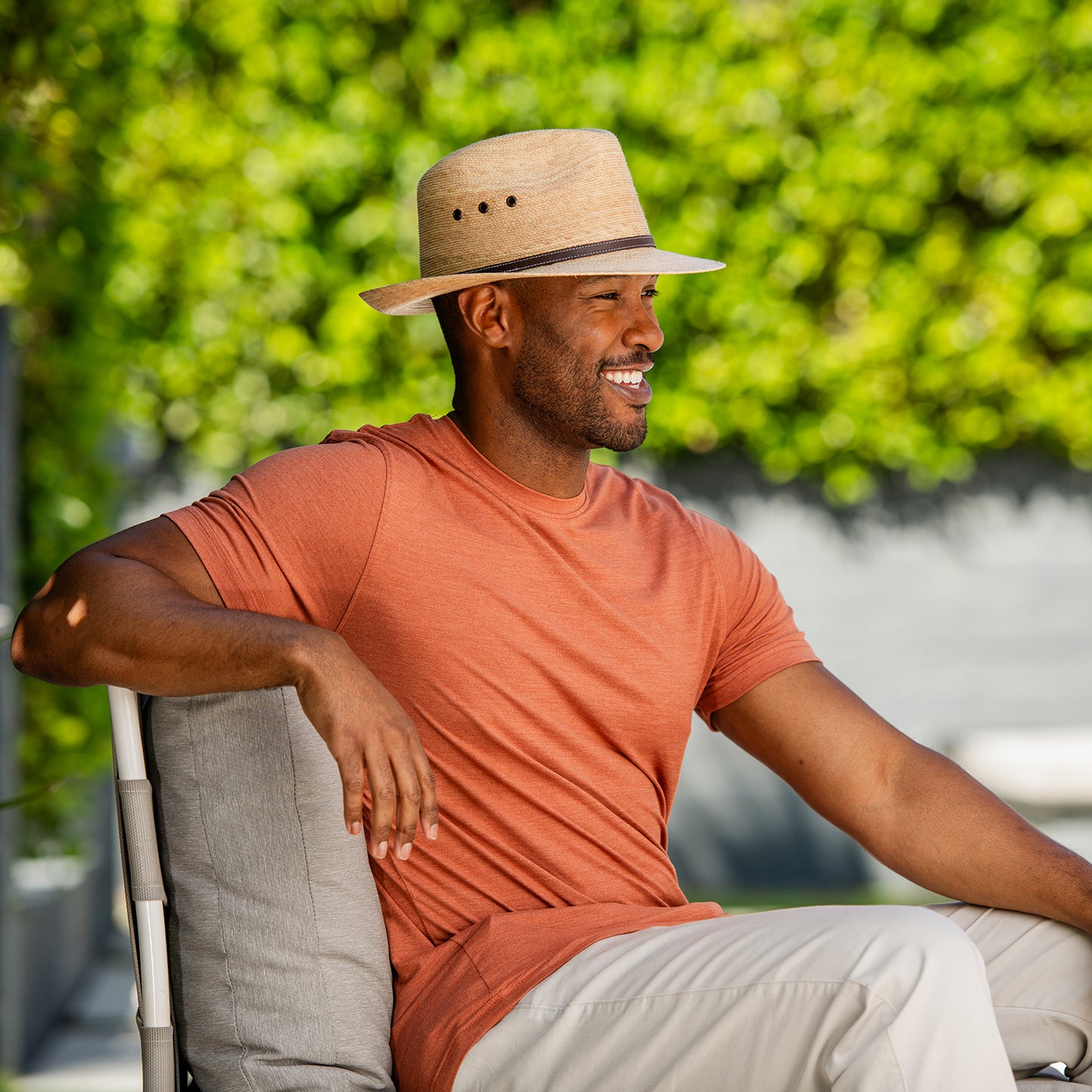Man wearing a palm fiber Cortez hat and orange shirt sitting outdoors with greenery in the background, Camel