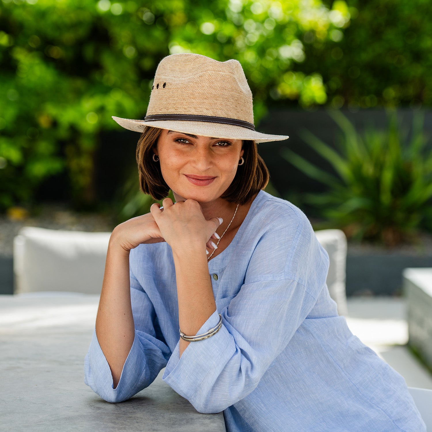 Woman wearing a Cortez trilby palm fiber sun protection hat and light blue outfit sitting outdoors with greenery in the background, Camel