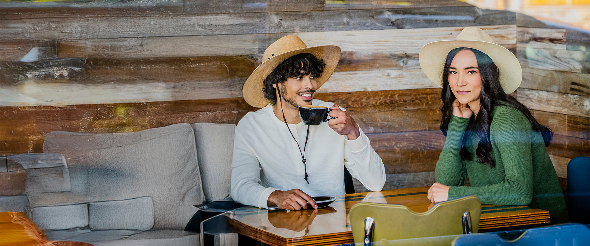 Two people sitting at a table with a wooden wall background, wearing the Baja and Tulum artisanal hats.