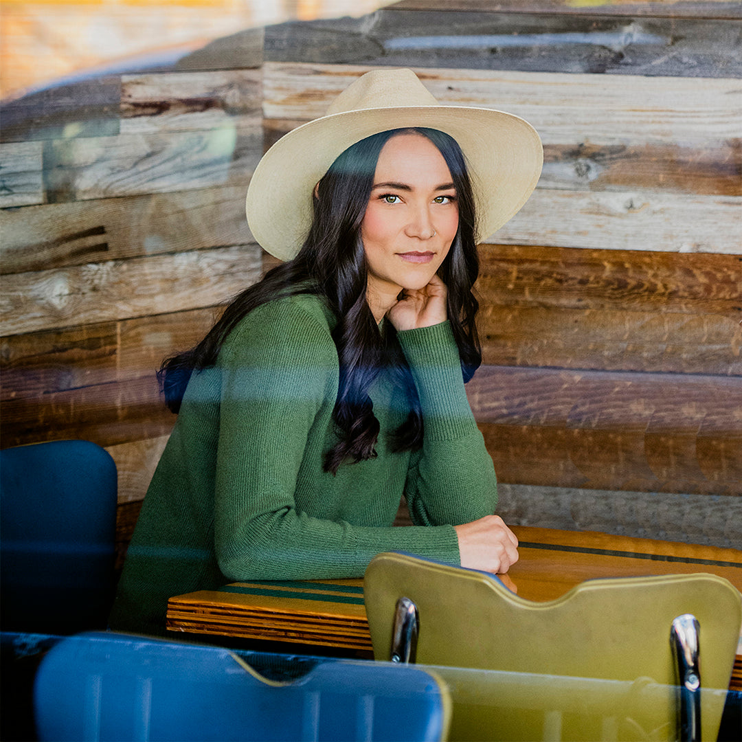 Woman wearing a Tulum artisanal hat and green sweater sitting in front of a wooden wall.