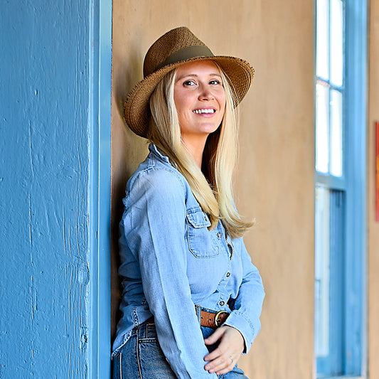 Woman wearing Carkella Emery fedora and denim shirt leaning against a blue and brown wall.