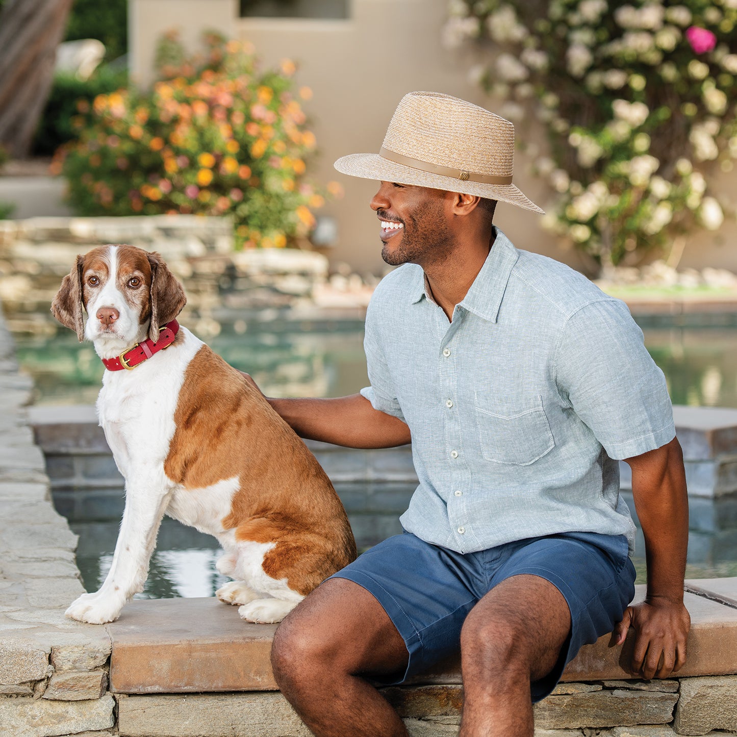 Man sitting with a dog by a poolside, surrounded by flowers, wearing men's Everett bamboo fedora travel sun hat, Mixed Camel