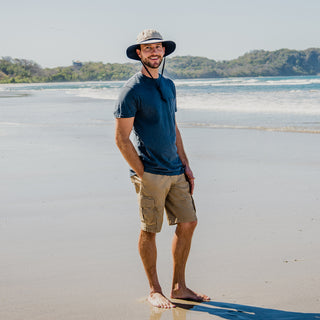 Man walking along a sunny beach wearing the Wallaroo Explorer Bucket Hat, blending wide-brim coverage and UPF 50+ protection for seaside adventures, Camel/Navy