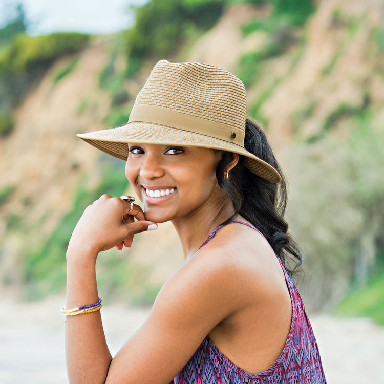 Woman enjoying a sunny beach day in the Wallaroo Gabi Wide Brim Sun Hat, designed with a ponytail-friendly opening, packable material, and wide-brim sun protection for a carefree, active lifestyle, Beige