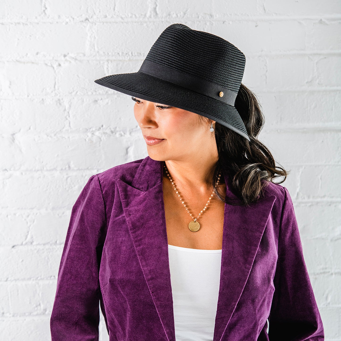 Woman wearing a black Gabi ponytail fedora hat and purple blazer against a white brick wall