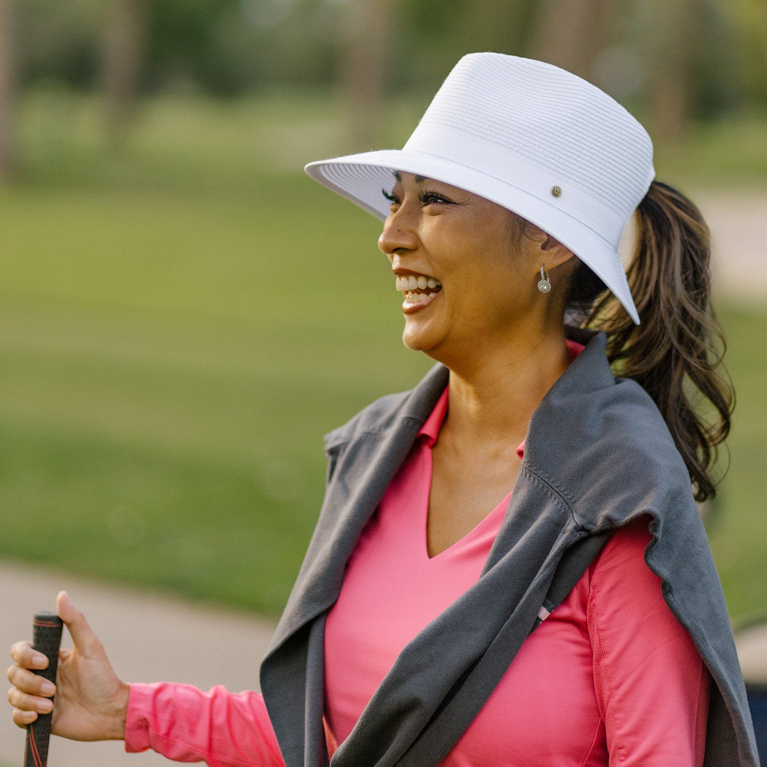 Woman in pink shirt and Gabi ponytail sun hat holding a golf club outdoors