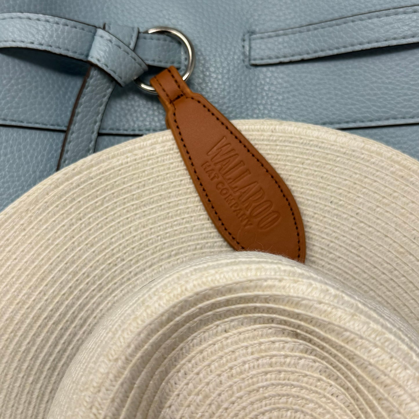Beige straw hat attached to blue bag with a brown leather hat clip 