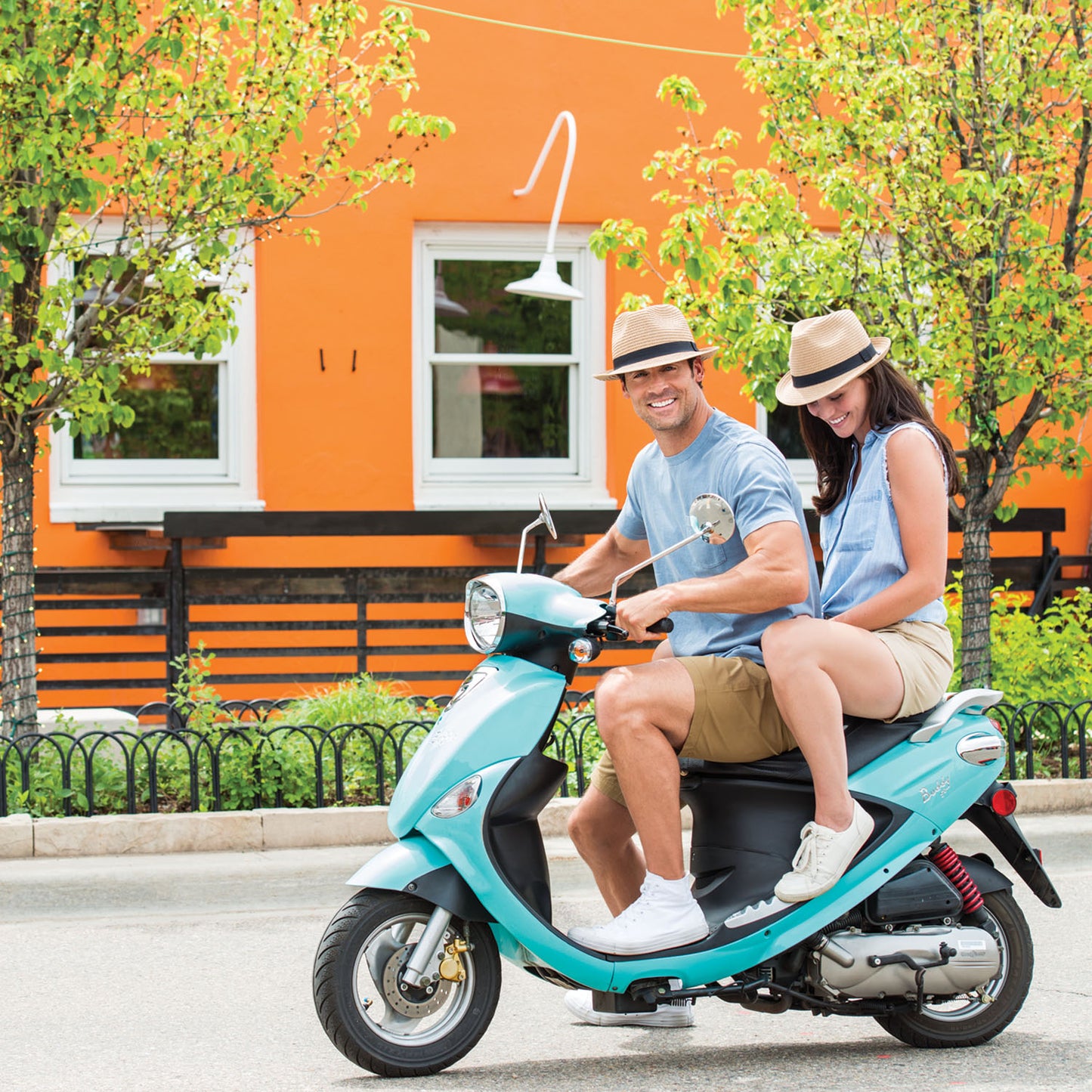 Friends riding a scooter across town, wearing the Wallaroo Justin Trilby Panama Hat for lightweight, short brim UPF 50+ sun coverage during active outings, Natural