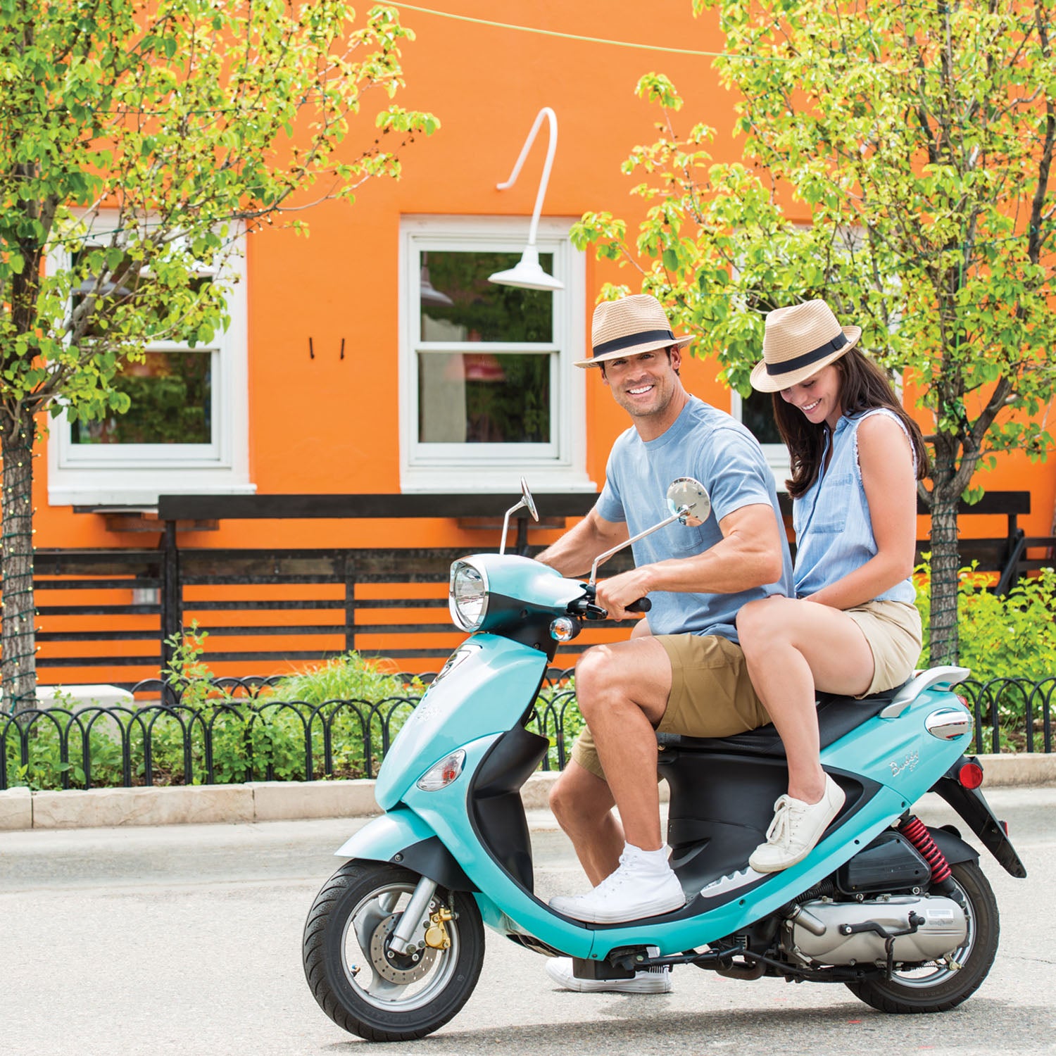 Friends riding a scooter across town, wearing the Wallaroo Justin Trilby Panama Hat for lightweight, short brim UPF 50+ sun coverage during active outings, Natural