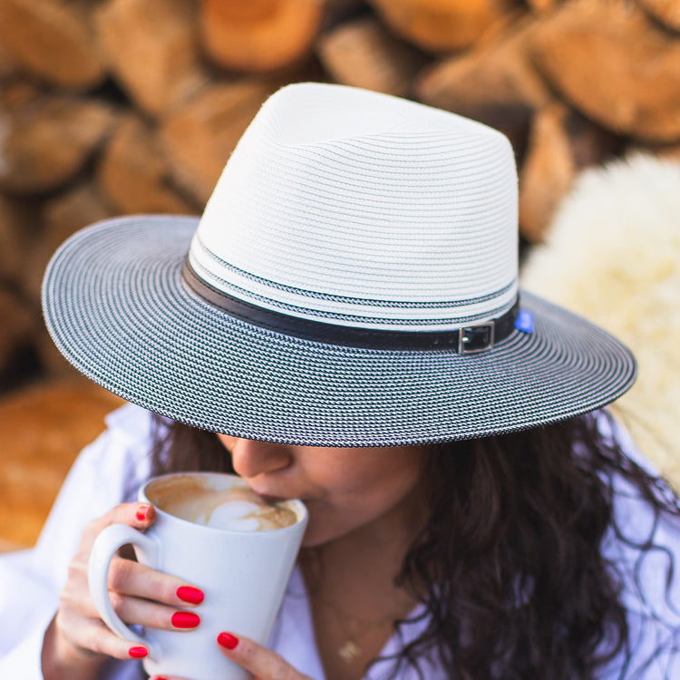 Woman enjoying a coffee at a cozy outdoor cafe, wearing the Wallaroo Women's Kristy Fedora Sun Hat with UPF 50+ protection, perfect for sunny days and urban outings, Ivory/Black