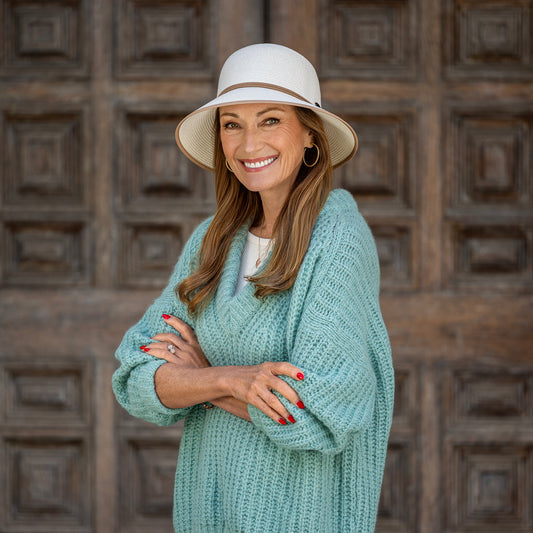 Actress Jane Seymour wearing the Lady Brett UPF 50+ cloche sun hat in ivory and a chunky mint green knit cardigan stands with arms crossed in front of a dark brown wooden door.