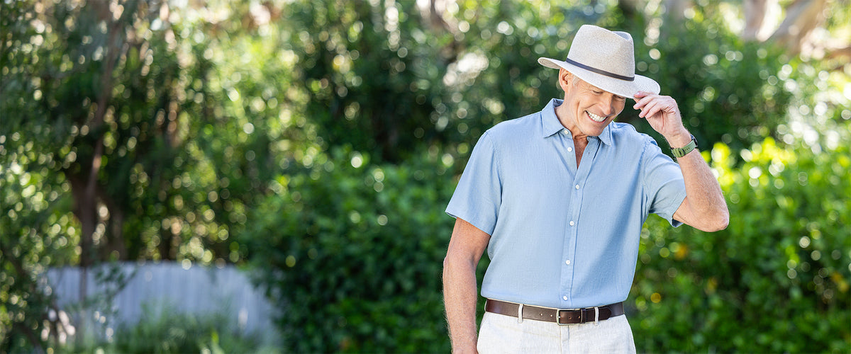 A man standing outdoors wearing the Finn UPF 50+ packable hat in mixed camel, a light blue short-sleeve button-up shirt, and white trousers, with a blurred green garden background.