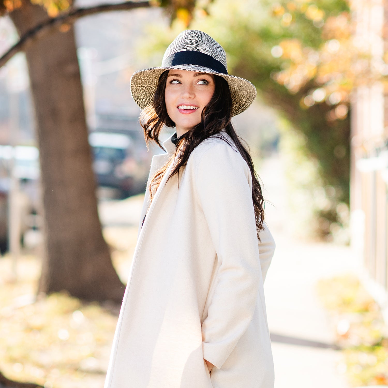 Woman wearing a white coat and Carkella Newhall hat in an outdoor setting with trees and blurred background