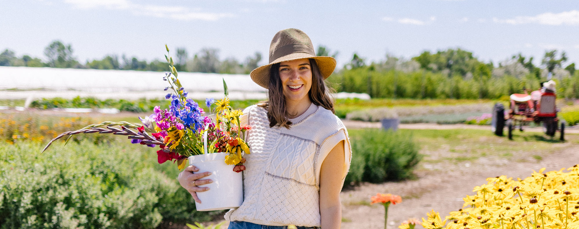 Woman wearing Carkella by Wallaroo Emery fedora holding a bouquet of flowers in a field with a tractor in the background