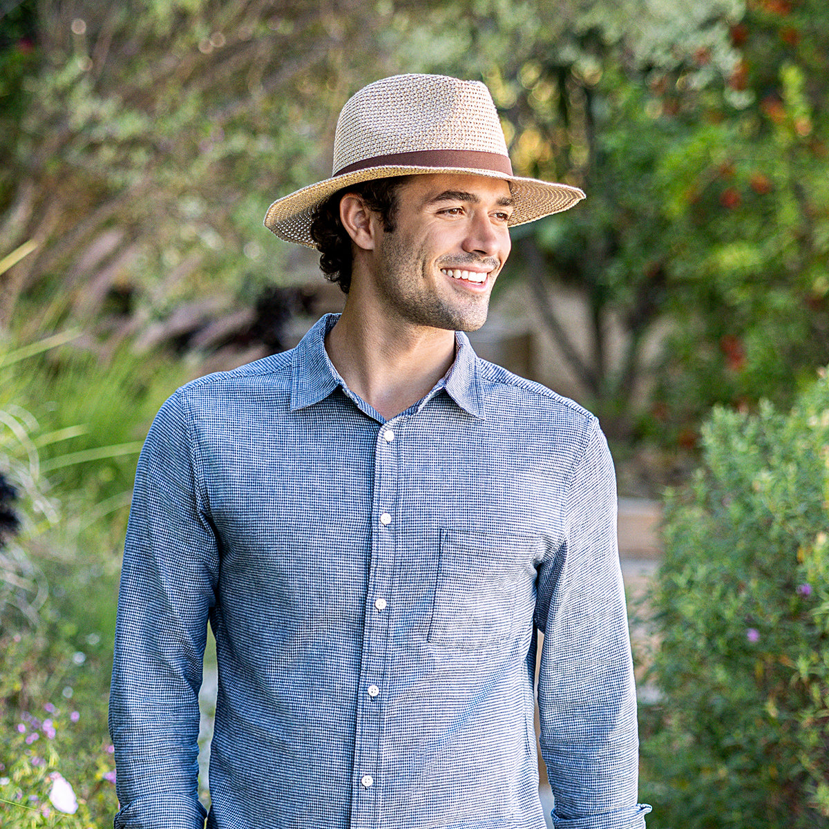 A man wearing the UPF 50+ packable Outback fedora hat in natural and a blue textured long-sleeve button-up shirt, standing outdoors in a garden setting.