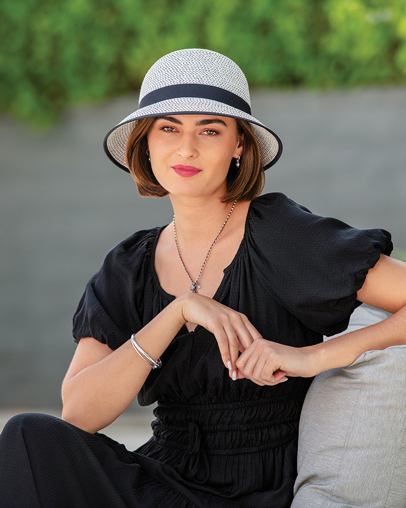 Woman wearing a black dress and Petite Darby sun hat with a black band, sitting outdoors.