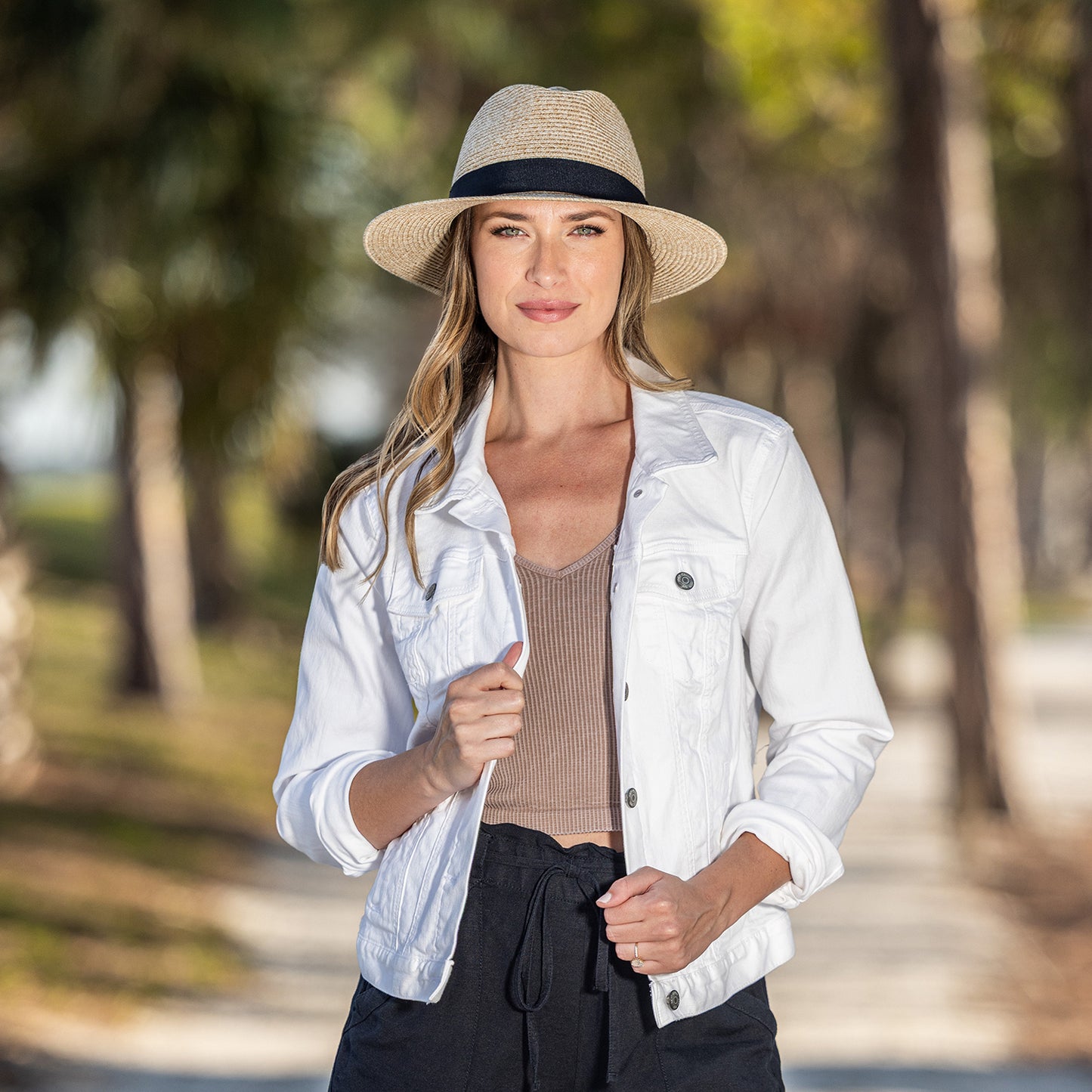 Woman wearing a white jacket, brown top, and Petite Palm Beach UPF 50+ packable fedora in front of trees.