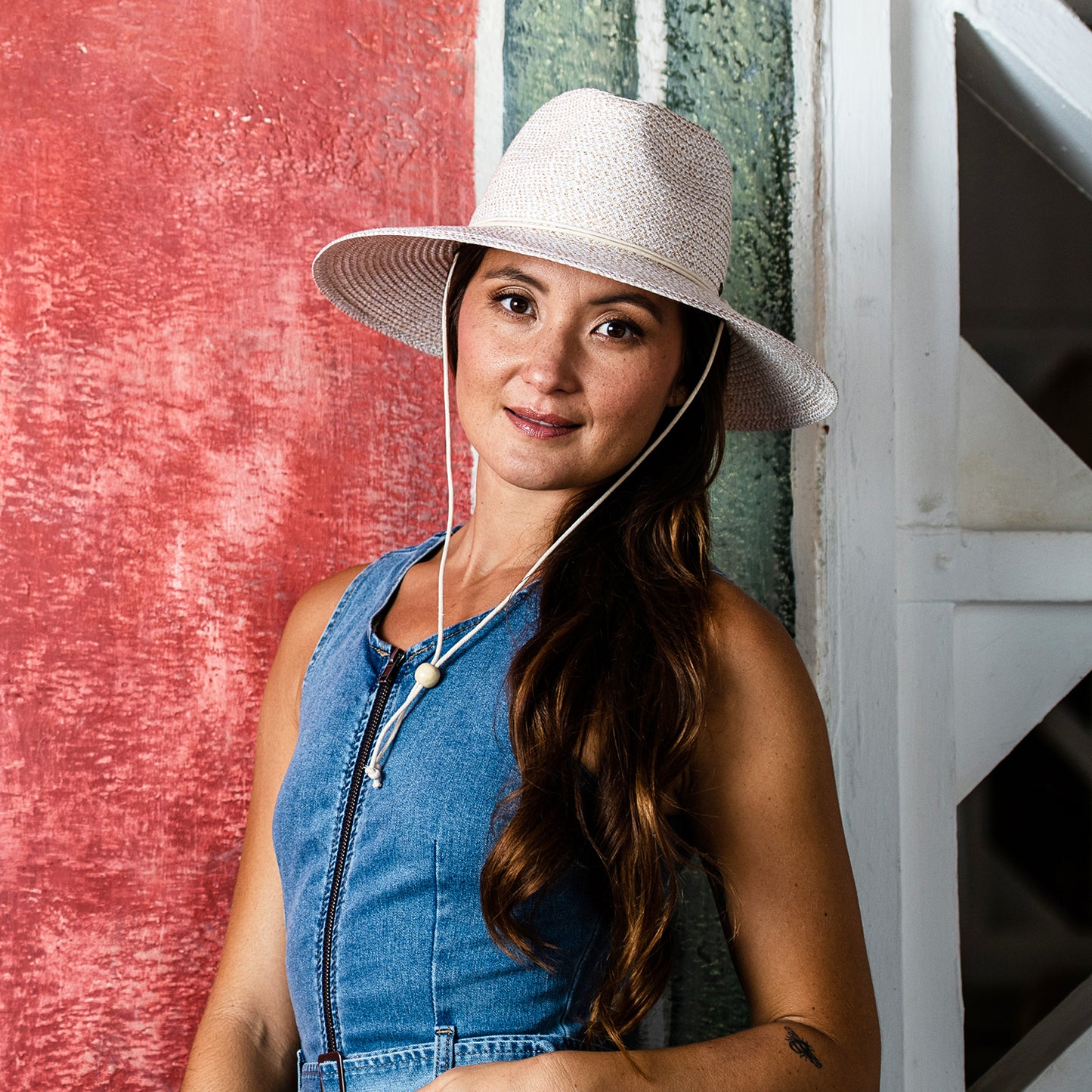 Woman wearing a wide-brimmed Petite Sanibel sun hat and denim dress against a textured red and green wall.