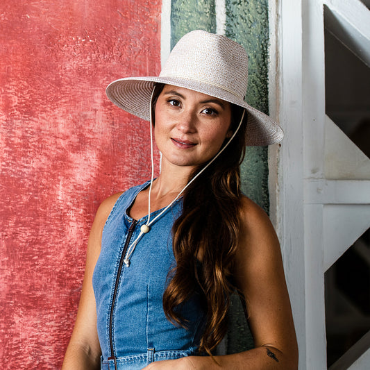 Woman wearing a wide-brimmed Petite Sanibel sun hat and denim dress against a textured red and green wall.