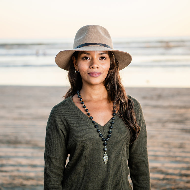 Woman bundled up on a fall day near water, wearing the Wallaroo Petite Aspen wide brim sun hat, designed to provide sun protection and style in cooler, natural landscapes, Camel