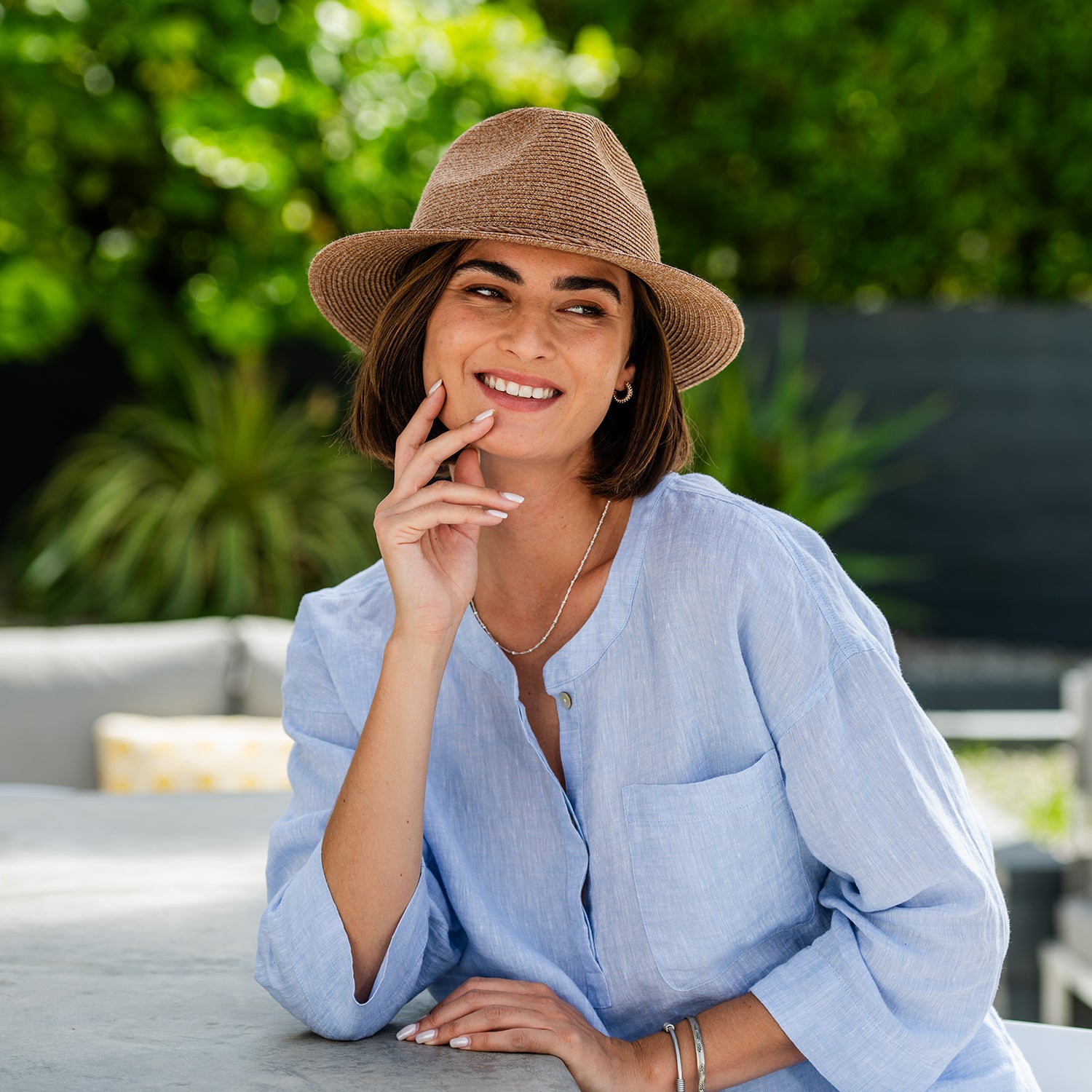 Woman wearing a Rio fedora sun hat and light blue shirt sitting outdoors with greenery in the background, Cinnamon