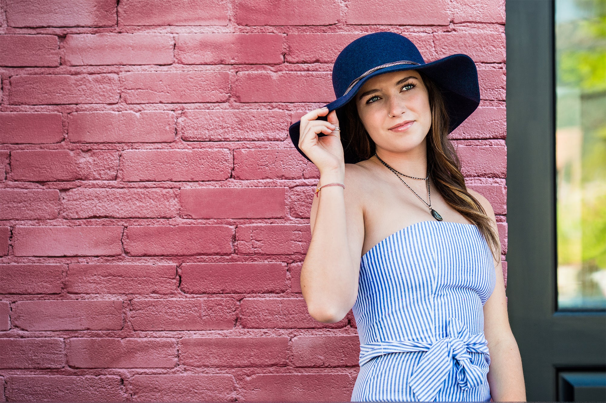 A woman wearing a blue and white striped strapless dress and a Cambria navy blue wide-brim hat, posing against a pink brick wall.
