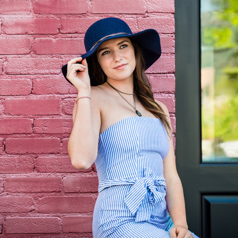 A woman wearing a blue and white striped strapless dress and a Cambria navy blue wide-brim hat, posing against a pink brick wall.
