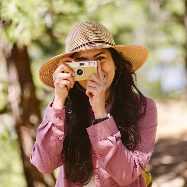 Woman taking photos wearing the Wallaroo Women’s Sanibel Sun Hat, a wide-brimmed UPF 50+ fedora with a chin strap, ideal for outdoor adventure, Camel