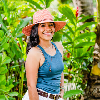 Woman in a tropical resort garden wearing the Wallaroo Sanibel Sun Hat, a fedora with UPF 50+ sun protection and a secure chin strap for breezy settings, Coral