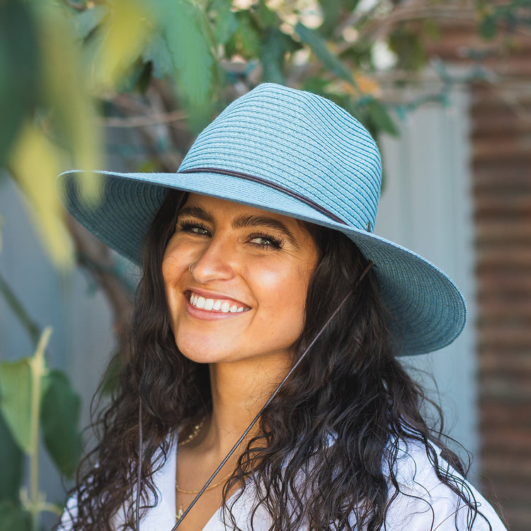 Woman protected from the sun in the Wallaroo Women’s Sanibel Fedora, with a wide brim and chin strap for windy summer escapes, Cornflower