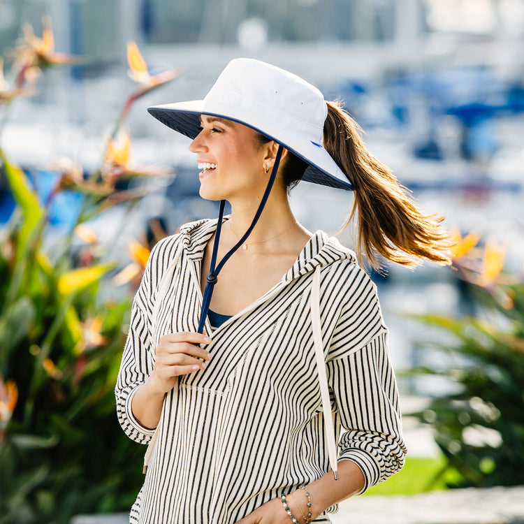 Woman at a tropical resort, shaded by the Wallaroo Sanitas Bucket Hat, featuring a wide brim and chin strap for breezy summer settings, White/Navy