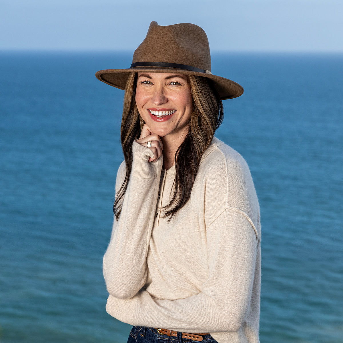 A woman smiling outdoors by the ocean, wearing a cream knit sweater and a Wallaroo UPF 50+ Snowmass fedora sun hat in camel.
