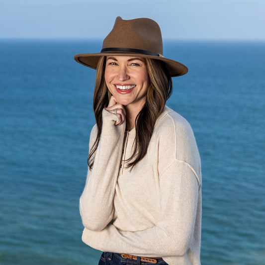 A woman smiling outdoors by the ocean, wearing a cream knit sweater and a Wallaroo UPF 50+ Snowmass fedora sun hat in camel.