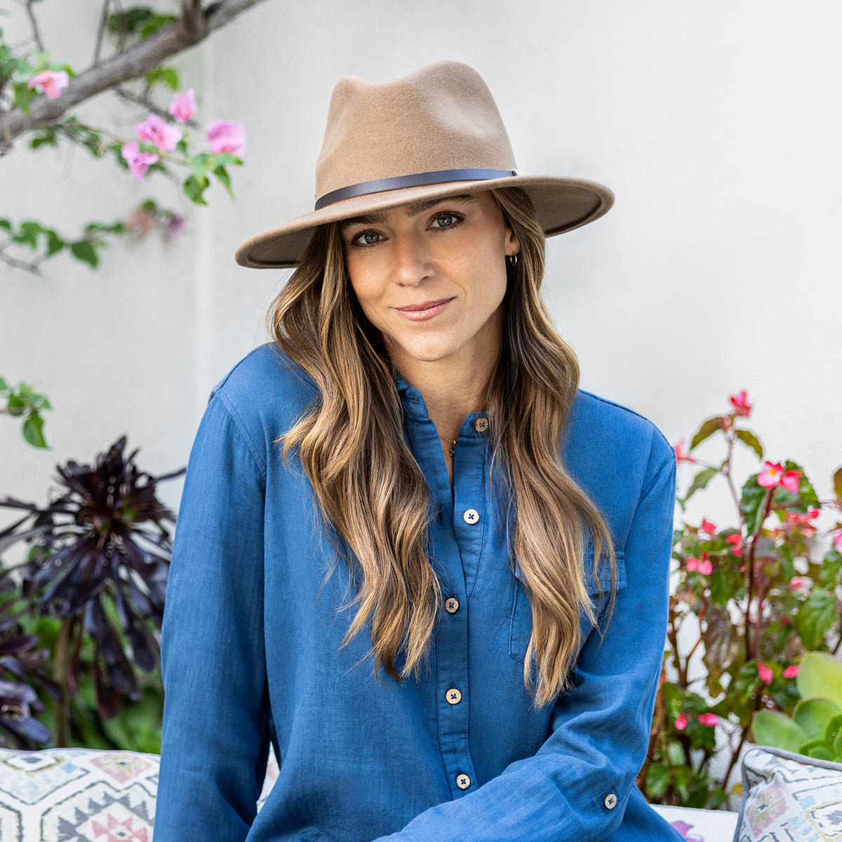 A woman wearing the Wallaroo UPF 50+ Snowmass fedora sun hat in camel in a blue long-sleeve button-up shirt, seated outdoors against a white wall with green plants and pink flowers.